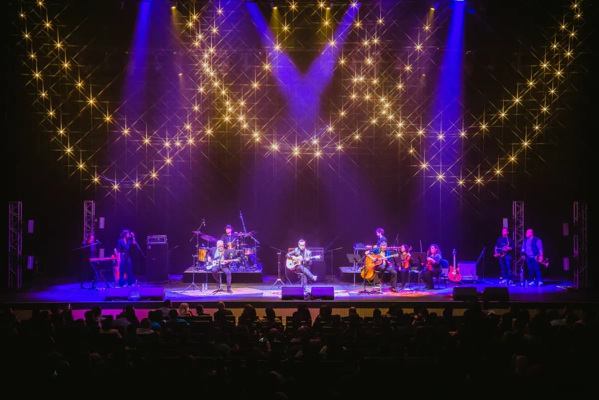 Full band performing acoustic set on theater stage with twinkling lights and purple-blue lighting