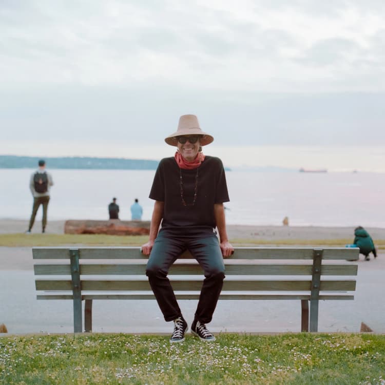 Clemente Castillo sitting on a waterfront bench wearing a straw hat and sunglasses with ships visible in the background